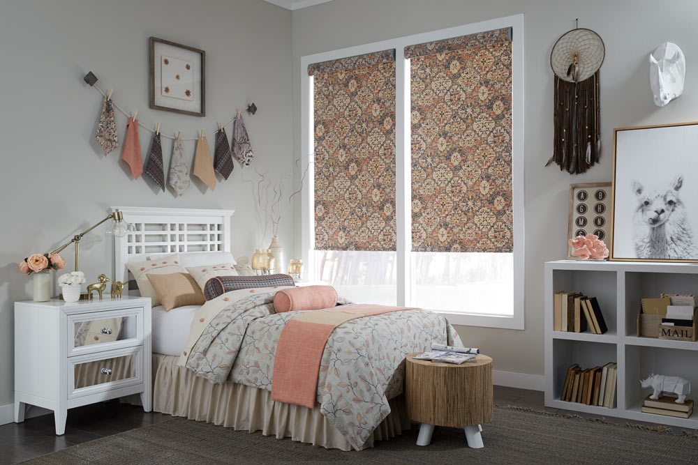 closeup of a bed with pink, purple, light pink, tan, and white Interior Masterpieces® Pillows and matching Custom Bedding with a window that has Fabric Shades that match