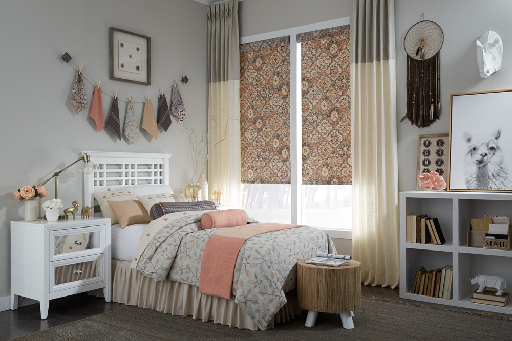 closeup of a bed with pink, purple, light pink, tan, and white Interior Masterpieces® Pillows and matching Custom Bedding with a window that has Draperies and Fabric Shades that match