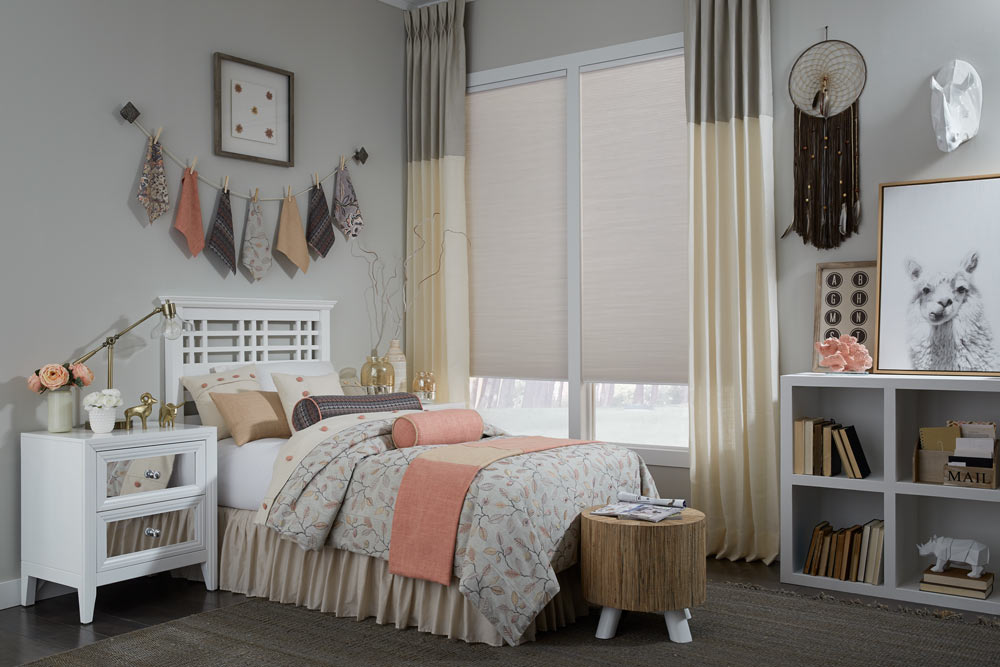 closeup of a bed with pink, purple, light pink, tan, and white Interior Masterpieces® Pillows and matching Custom Bedding with a window that has Draperies and Fabric Shades that match