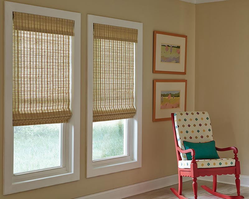 Manh Truc® Woven Wood Shades Three cornsilk colored child-safe flat roman shades hang in a tan room with a red rocking chair.