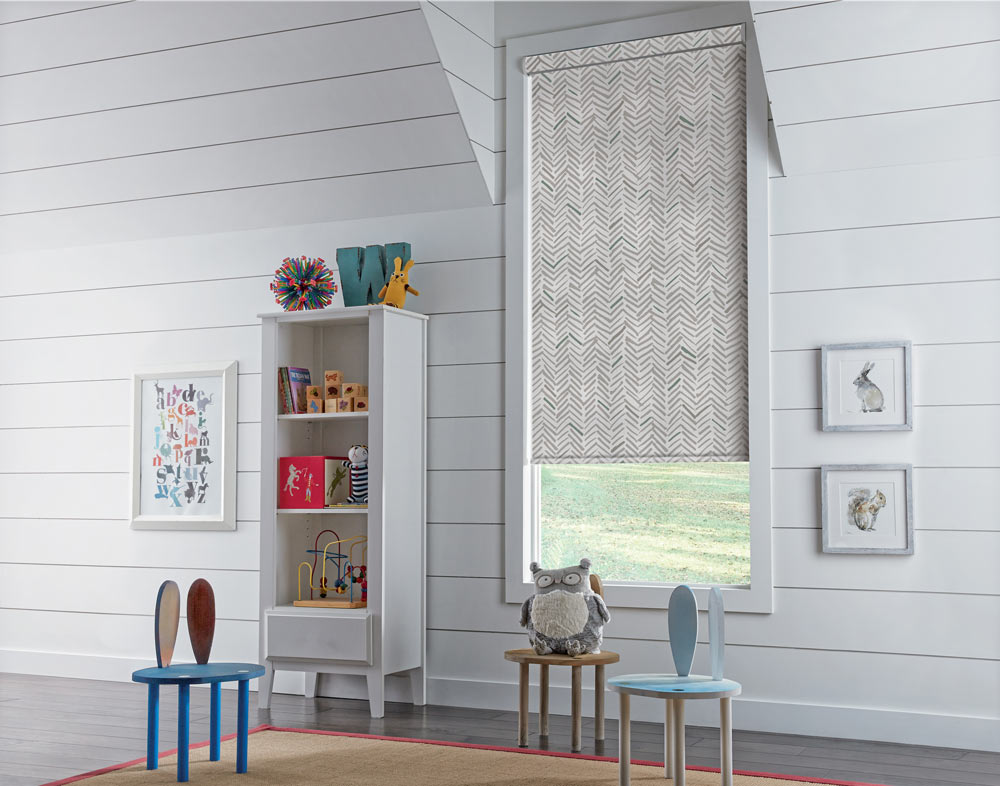 a Genesis® Roller Shade with a green and gray chevron patterned shade against white walls in a kid's room with little chairs and a stuffed owl in the foreground