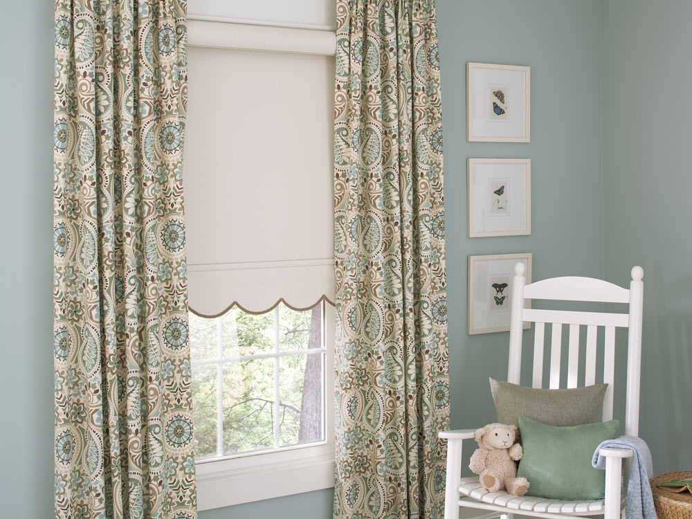 off white Genesis® Cassette Roller Shade with scallop and brown Embellishment Trim and tan, light green, and brown Draperies on either side next to a white chair with custom pillows and a stuffed sheep on it