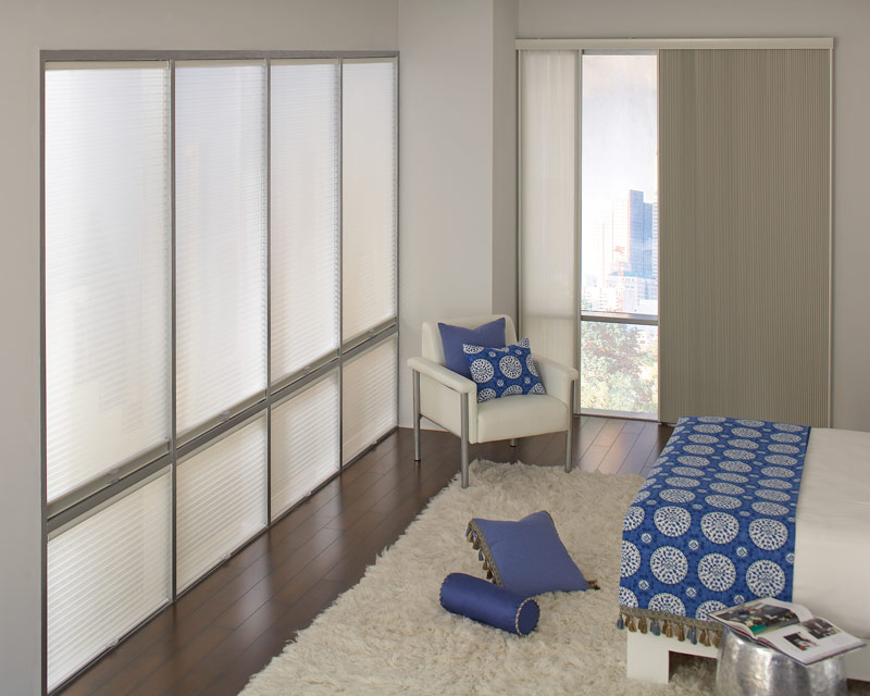 Parasol® Cellular Shades several large tan Parasol® Cellular Shades on one wall and a large Glissade sliding panel system on the other in a room with a bed that has blue Interior Masterpieces® bedding and accessories on it